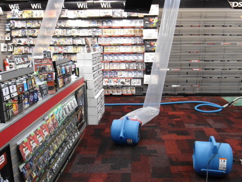 An empty video game store with Wii and PS3 games on shelves, a blue industrial carpet cleaner in the center for water damage restoration, and two blue chairs on the right.