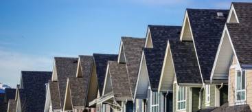 A row of similar suburban houses with pointed roofs and varying pastel-colored siding under a clear blue sky, some showing signs of water damage restoration.